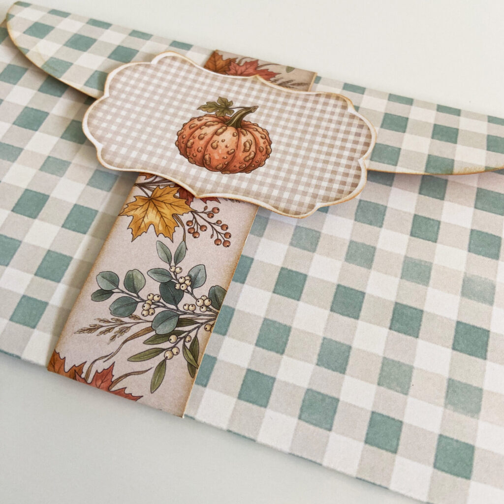 Close up of a Pumpkin Conservatory folio folded and closed, wrapped with a belly band and badge, in sage green gingham, and adorned with autumn leaves and a pumpkin.
