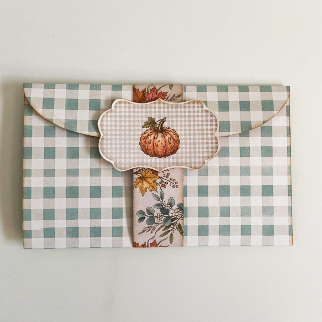 Pumpkin Conservatory folio folded and closed, wrapped with a belly band and badge, in sage green gingham, and adorned with autumn leaves and a pumpkin.